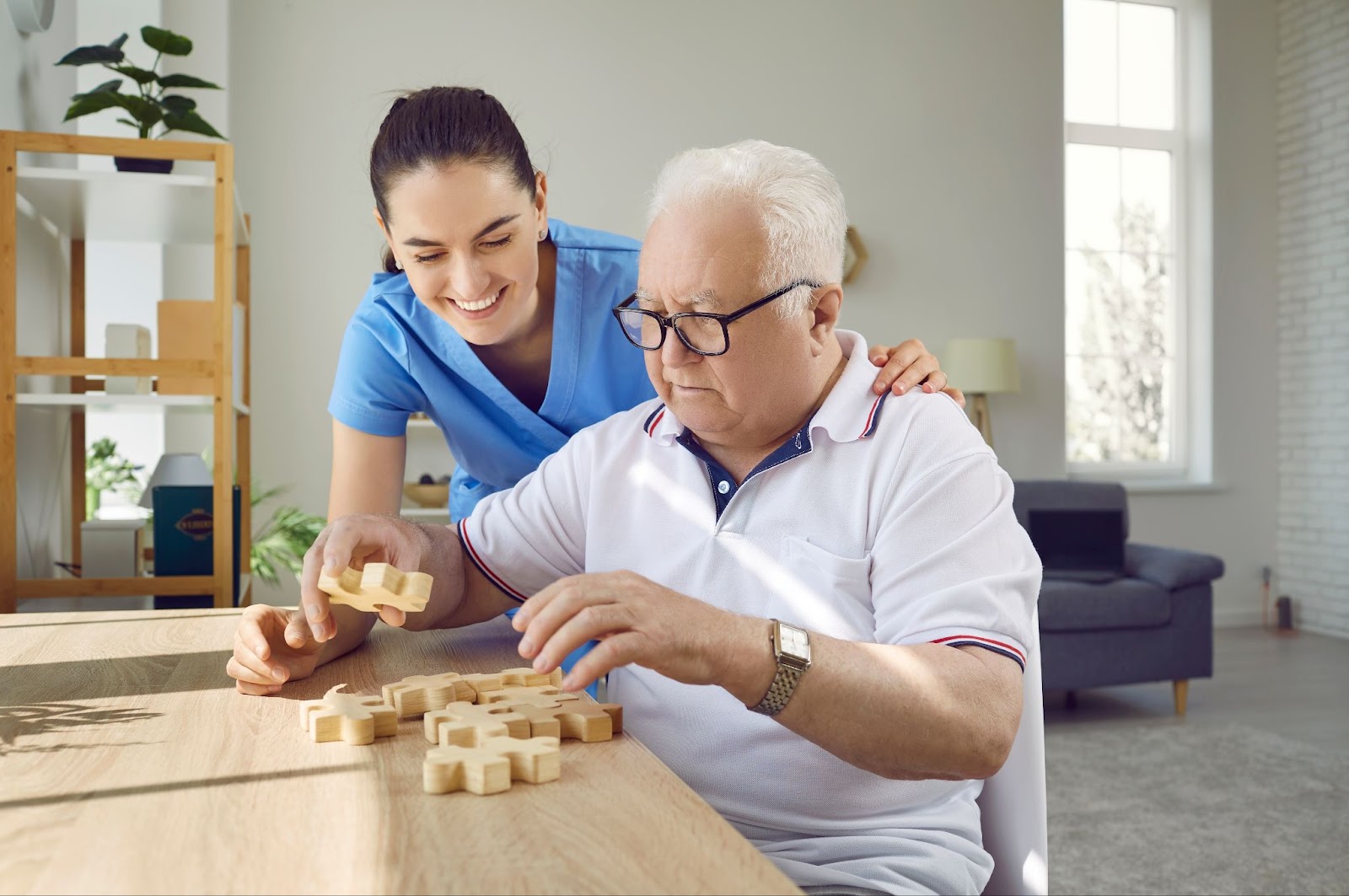 A resident and a caregiver sit at a table in the activity room of a care community, putting a puzzle together. The resident is deeply focused on the puzzle, and the caregiver smiles in encouragement.