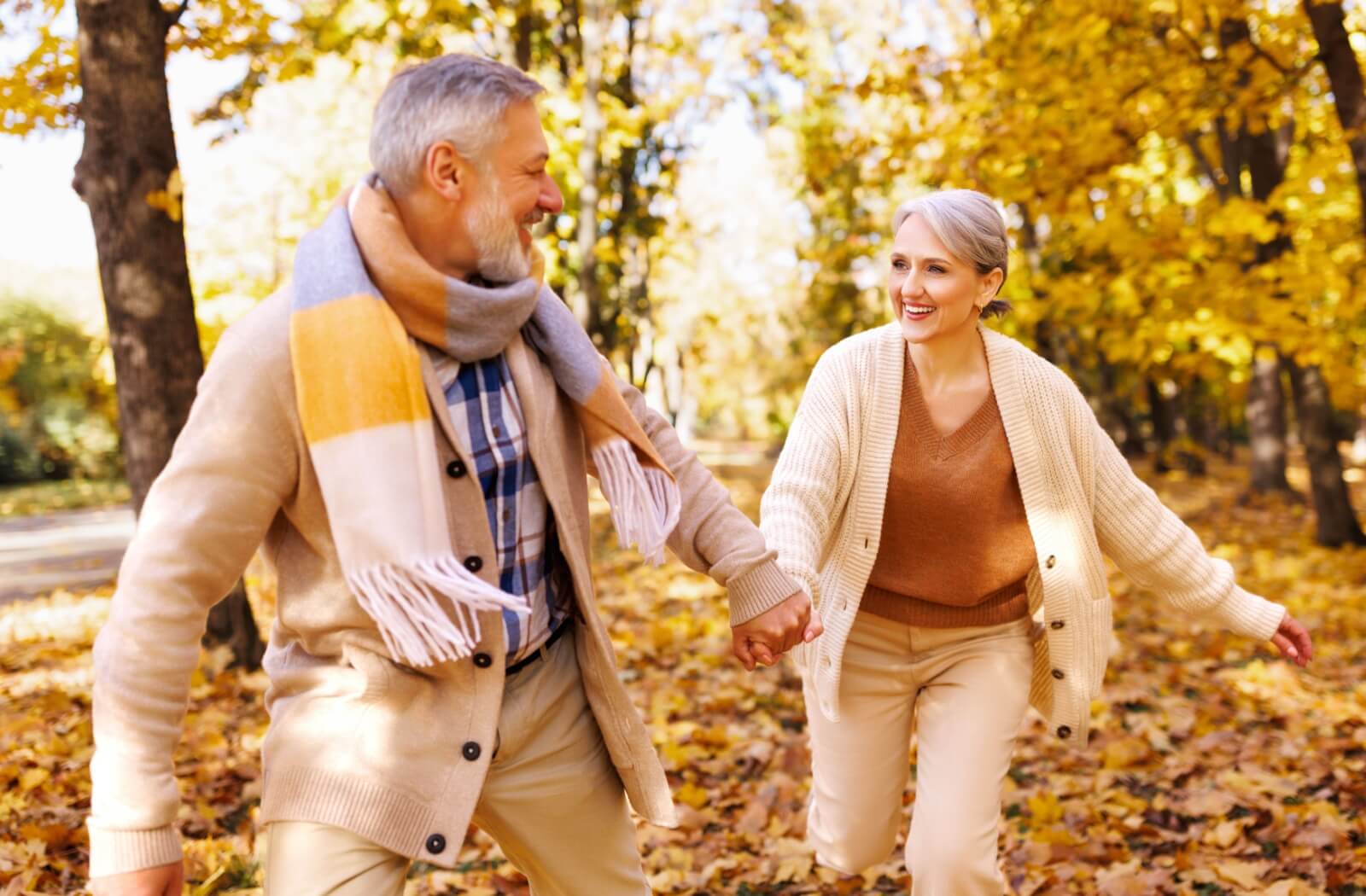 A senior couple wearing beige clothes hold hands while running through an outdoor pathway of fallen autumn leaves