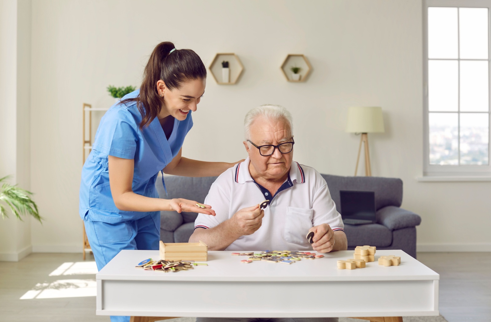 A caregiver in scrubs smiling and assisting an older adult in glasses with a puzzle activity at a table in a well-lit living space.