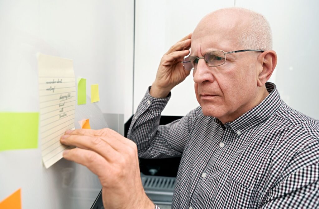 An older adult wearing glasses, concentrating while reading notes and post-its on a wall, appearing disoriented.