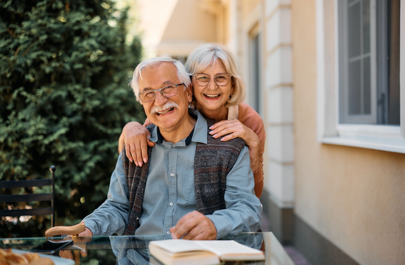 An older adult hugging their spouse from behind and smiling at the camera on the patio of their new retirement community.
