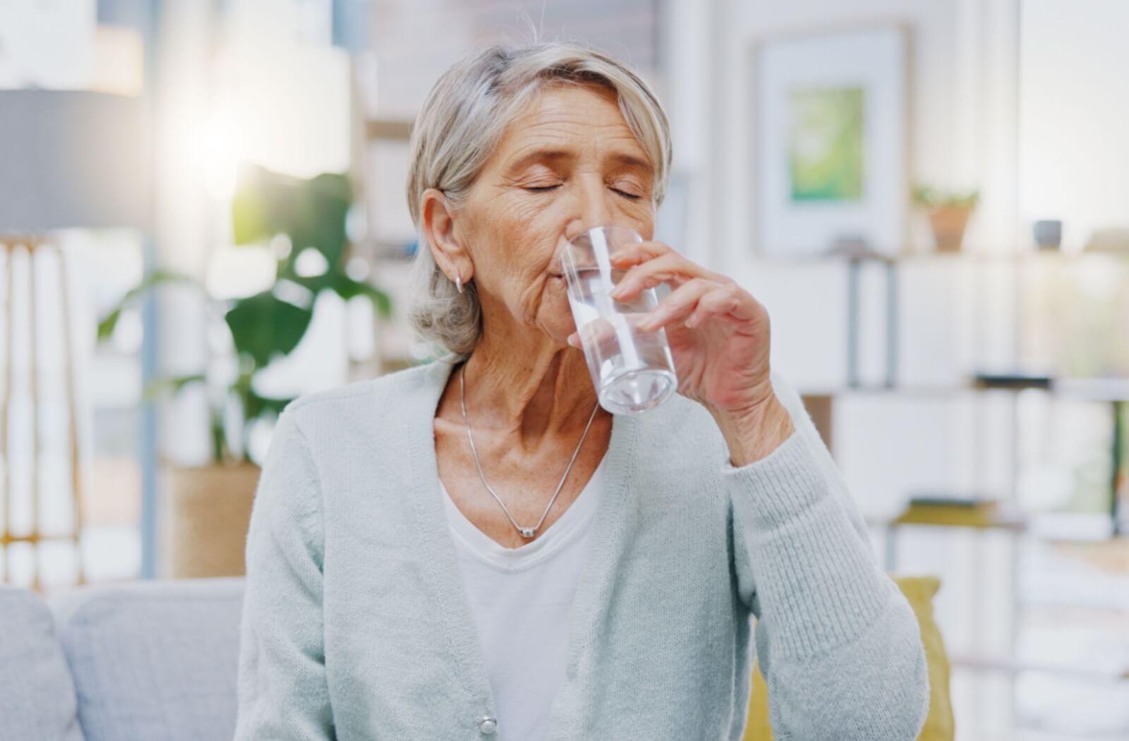 An older adult drinking a glass of water while sitting in their living room.