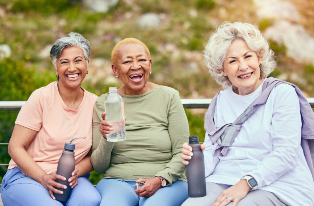 Three older adults sitting on a park bench and smiling while holding water bottles.