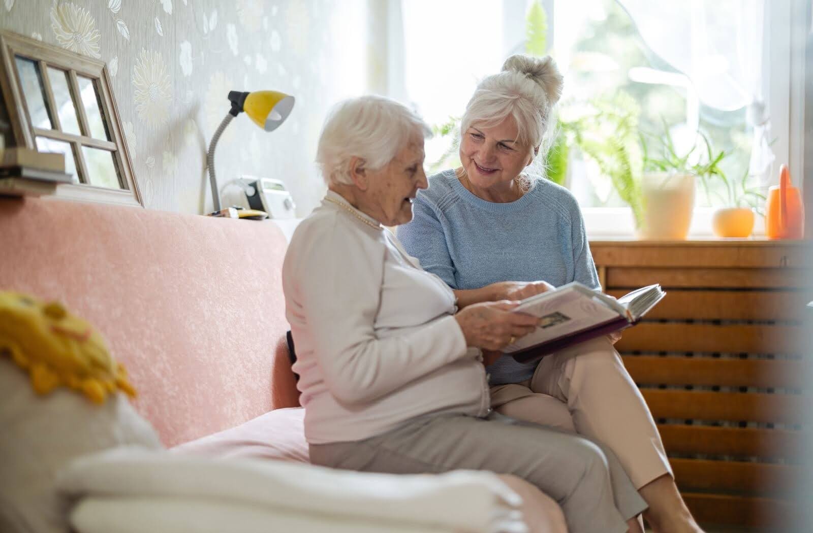 A senior living resident and her daughter smiling, as they share memories and look at a photo album in a cozy, sunlit living room.